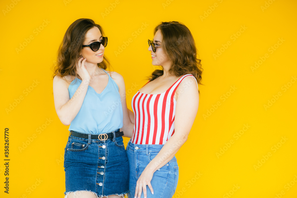 Two pretty excited young girls friends standing isolated over yellow background, pointing fingers at camera
