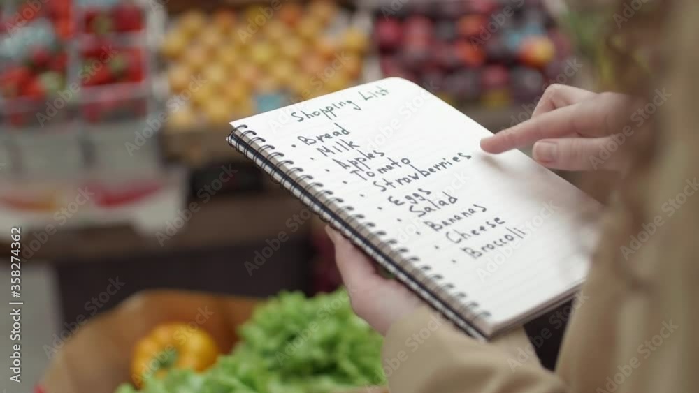 Rear view of unrecognizable woman standing in grocery marker, holding notepad with shopping list and analyzing it