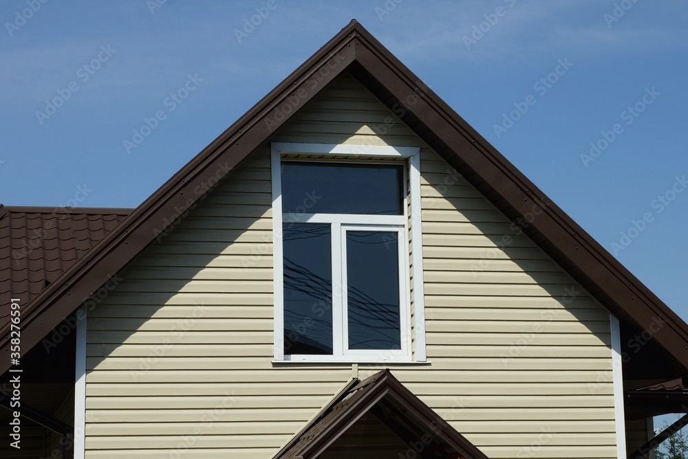 gray plastic attic of a private rural house with one window under a brown tiled roof against a blue sky