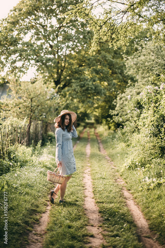 Young beautiful woman in a blue dress and hat with a basket in hand walks in the park on a summer trail. © lesia17