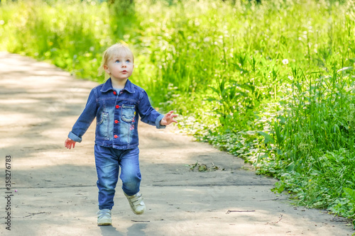 Selective focus on a blonde little girl running on an asphalt path in a park past thickets of dense green grass. Happy child plays in nature on a sunny day. The girl is dressed in a blue denim suit. © Nekrasov