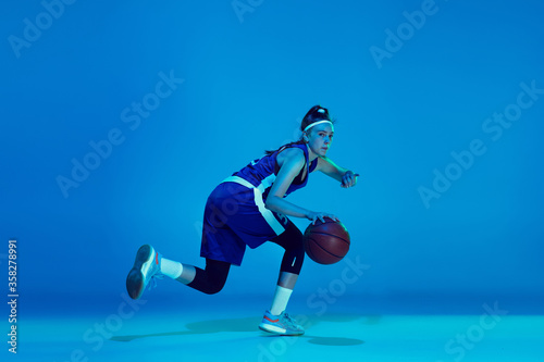 Goal achievement. Young caucasian female basketball player training, prcticing with ball isolated on blue background in neon light. Concept of sport, movement, energy and dynamic, healthy lifestyle.