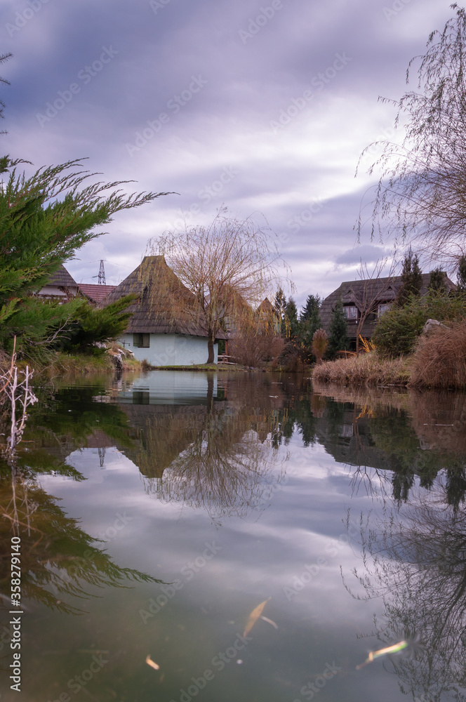 Obraz premium Old European style houses on the pond surrounded with threes and bushes. Moody sky and reflections in the pond. Vintage feel.