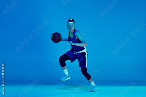 Goal achievement. Young caucasian female basketball player training, prcticing with ball isolated on blue background in neon light. Concept of sport, movement, energy and dynamic, healthy lifestyle.