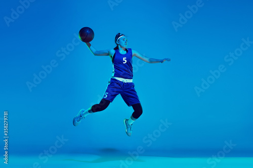 In high jump. Young caucasian female basketball player training, prcticing with ball isolated on blue background in neon light. Concept of sport, movement, energy and dynamic, healthy lifestyle.
