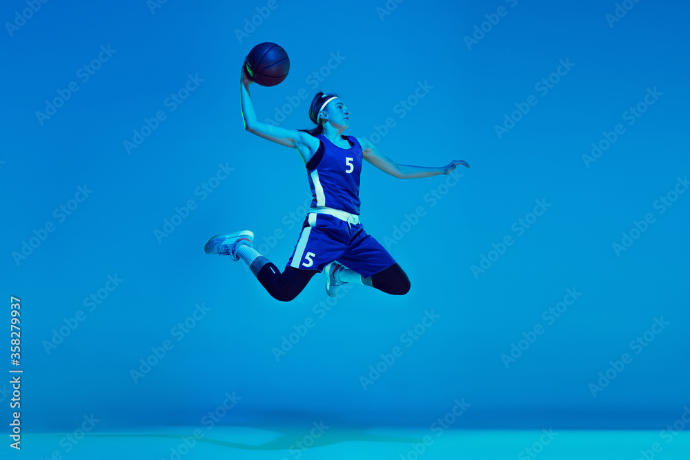 In high jump. Young caucasian female basketball player training, prcticing with ball isolated on blue background in neon light. Concept of sport, movement, energy and dynamic, healthy lifestyle.