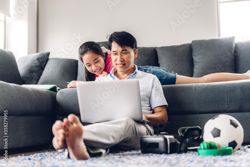 Smiling asian man relaxing using laptop computer working and video conference meeting chat with his girl daughter learning reading book and studying knowledge at home.work from home concept
