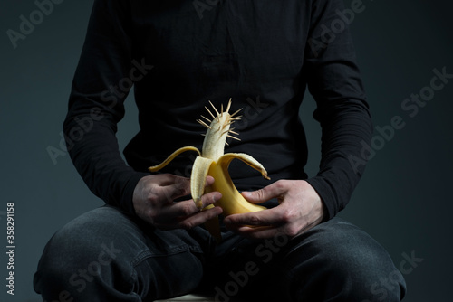 concept of genital cancer in men. A man in black holds a banana in his hands with thorns on his head, with a background on the male penis. On a gray background. photo