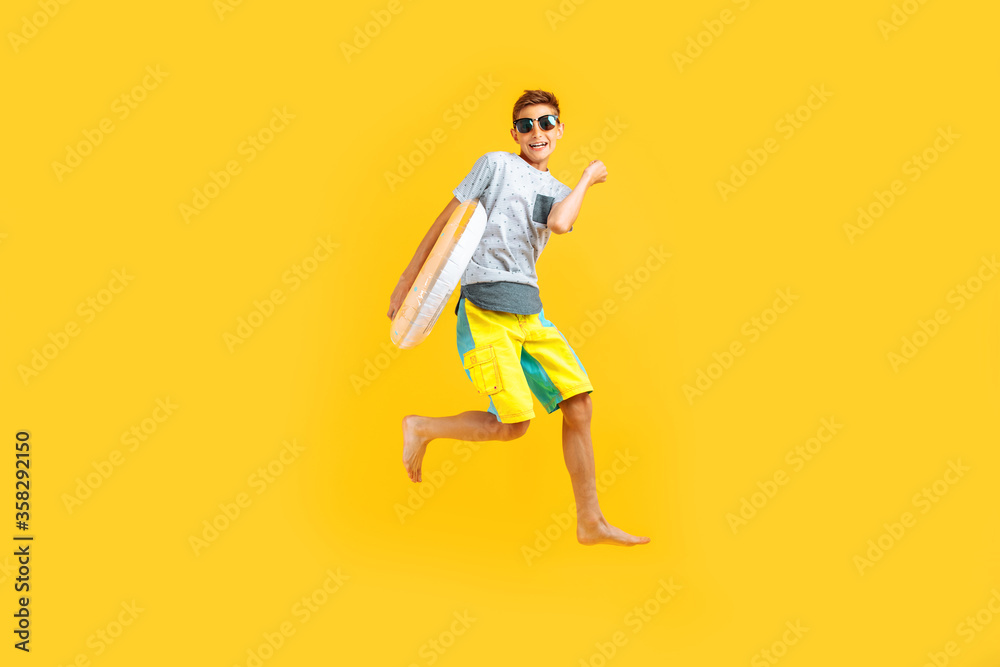happy excited teenager in sunglasses and a summer hat, having fun and jumping, holding an inflatable ring on a yellow background