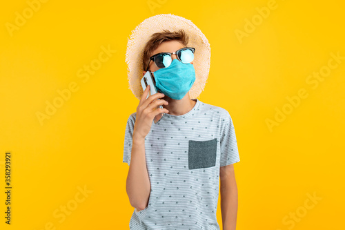 teenager in a summer hat and a medical protective mask on his face, talking on a mobile phone, on an yellow background