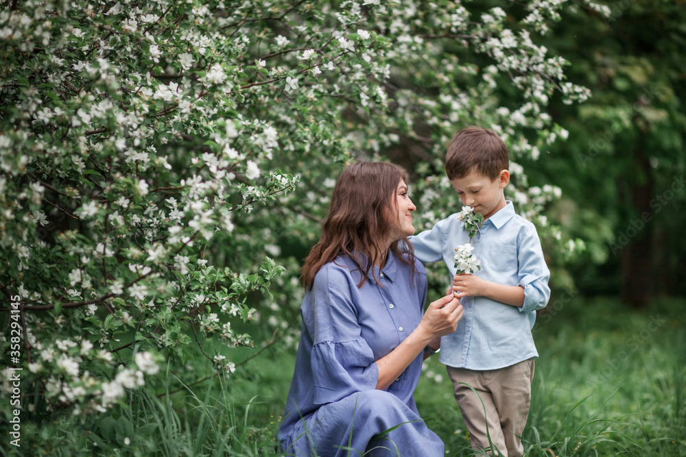 Fototapeta premium Happy and beautiful mother and son in clothes of blue tones are hugging next to a blossoming apple tree. The concept of spring, motherhood, happy family, relationships.
