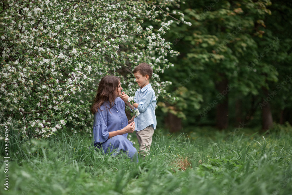 Happy and beautiful mother and son in clothes of blue tones are hugging next to a blossoming apple tree. The concept of spring, motherhood, happy family, relationships.