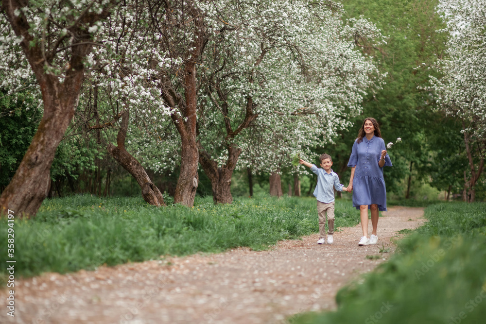 Fototapeta premium Happy and beautiful mother and son in clothes of blue tones walking in a blossoming apple orchard. The concept of spring, motherhood, happy family, relationships