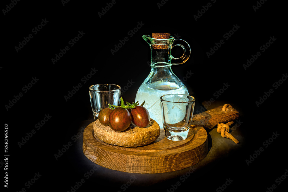 Two glasses and illuminated from the side decanter with vodka, raki or spirits, dry rye bread with cherry tomatoes on wooden board. Isolated on black background Two glasses and illuminated from the side decanter with vodka, raki or spirits, dry rye bread with cherry tomatoes on wooden board. Isolated on black background