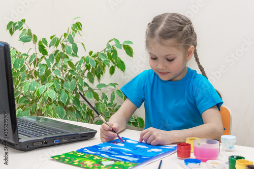 Distance learning online education. A sick preschooler in medical mask studies drawing from home. Girl does her homework, smiles and dips brush in the paint. Paint gouache, drawing on table.