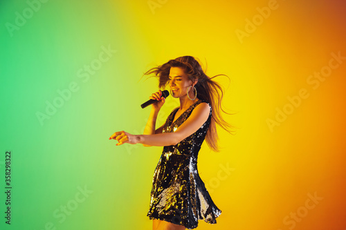 Caucasian female singer portrait isolated on gradient studio background in neon light. Beautiful female model in dress with microphone. Concept of human emotions, facial expression, ad, music, art.