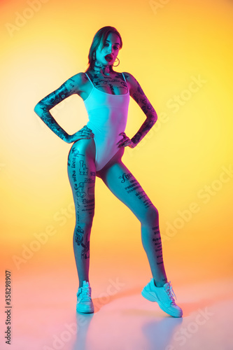 Beauty. Young caucasian woman in swimsuit posing sensual on gradient yellow background in neon. Beautiful model with tattoos. Human emotions, sales, ad concept. Resort and vacation, summertime.