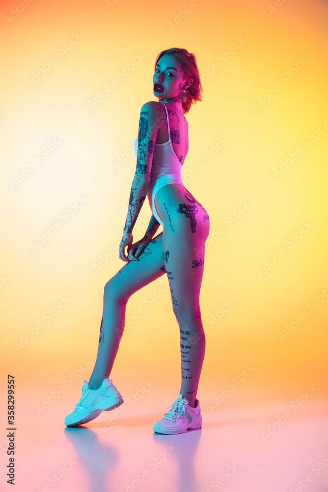 Fototapeta Fashion. Young caucasian woman in swimsuit posing sensual on gradient yellow background in neon. Beautiful model with tattoos. Human emotions, sales, ad concept. Resort and vacation, summertime.