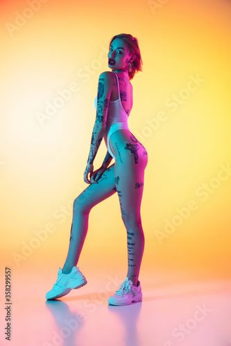 Fashion. Young caucasian woman in swimsuit posing sensual on gradient yellow background in neon. Beautiful model with tattoos. Human emotions, sales, ad concept. Resort and vacation, summertime.