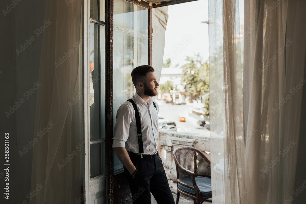 Young businessman standing at window looking at city scenery on the background. Motivation concept. Business trip. Businessman in luxury room of the expensive beautiful hotel. Copy space. Young businessman standing at window looking at city scenery on the background. Motivation concept. Business trip. Businessman in luxury room of the expensive beautiful hotel. Copy space.