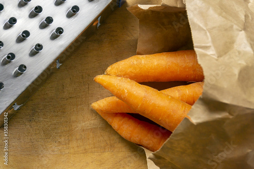 Fresh carrots in recyclable bag and metal grater on a wooden chopping board. Homemade recipe, cooking, food ingredient, kitchen utensils.