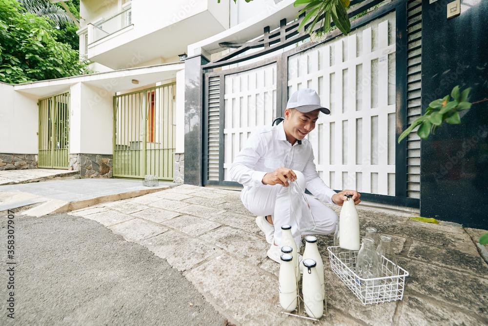 Joyful Asian milkman wearing perfect white uniform delivering milk and taking empty bottles back in early morning, copy space