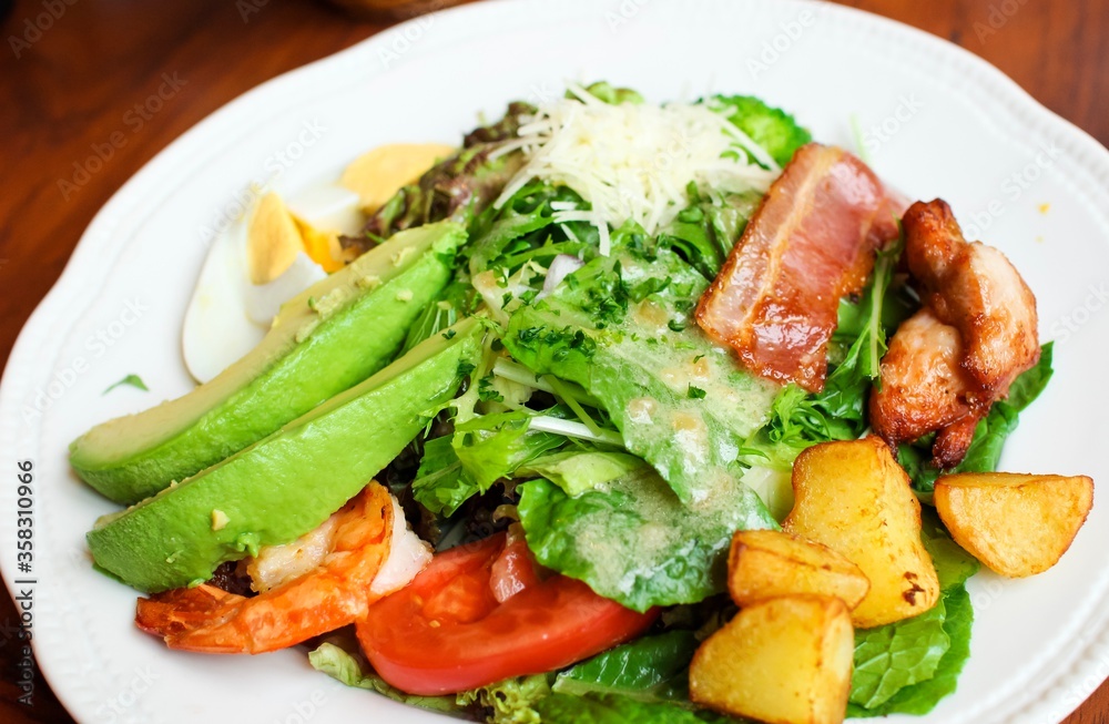 Colorful salad with tomatoes, avocado, lettuce, eggs, ham, chicken, potatoes and cheese on a white plate