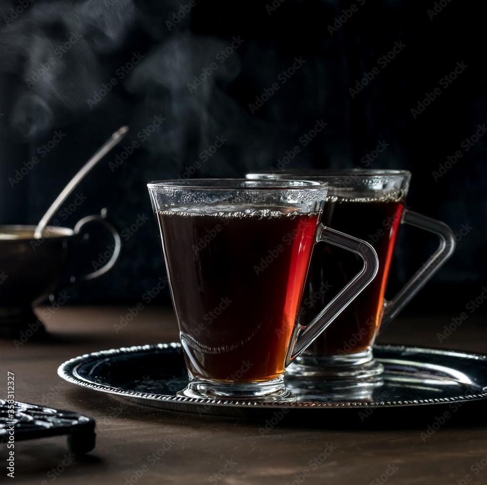 A close up view of two glasses of steaming hot tea on a metal tray against a black background. A close up view of two glasses of steaming hot tea on a metal tray against a black background.