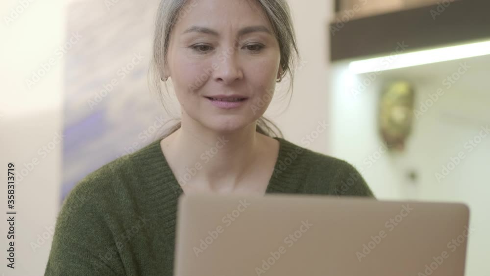 Pleased attractive mature woman with long gray hair using laptop computer while sitting by the table indoors