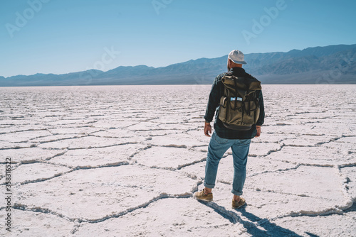 Back view of male traveler exploring wild nature environment of death valley with desolate salt surface, hipster guy wanderlust with backpack looking at horizon with high rocks in Badwater basin