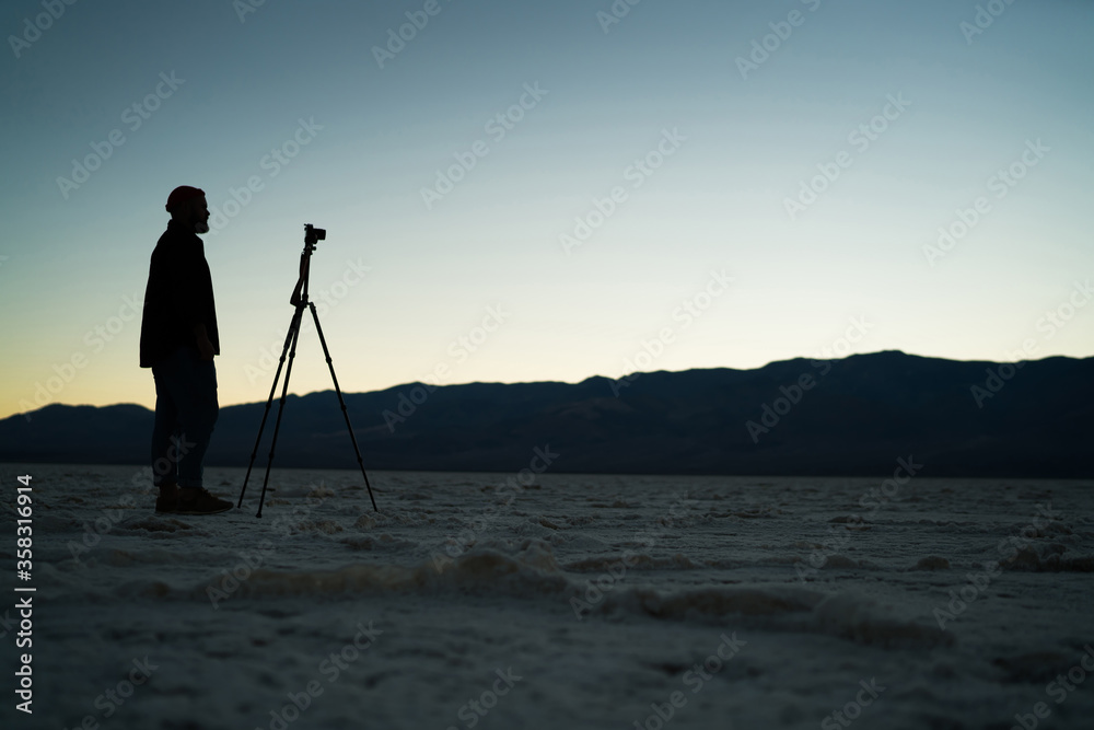 Male silhouette of photographer standing near tripod having expedition in death valley in twilight, man using professional equipment for making night shooting of wild nature and landscape in desert.