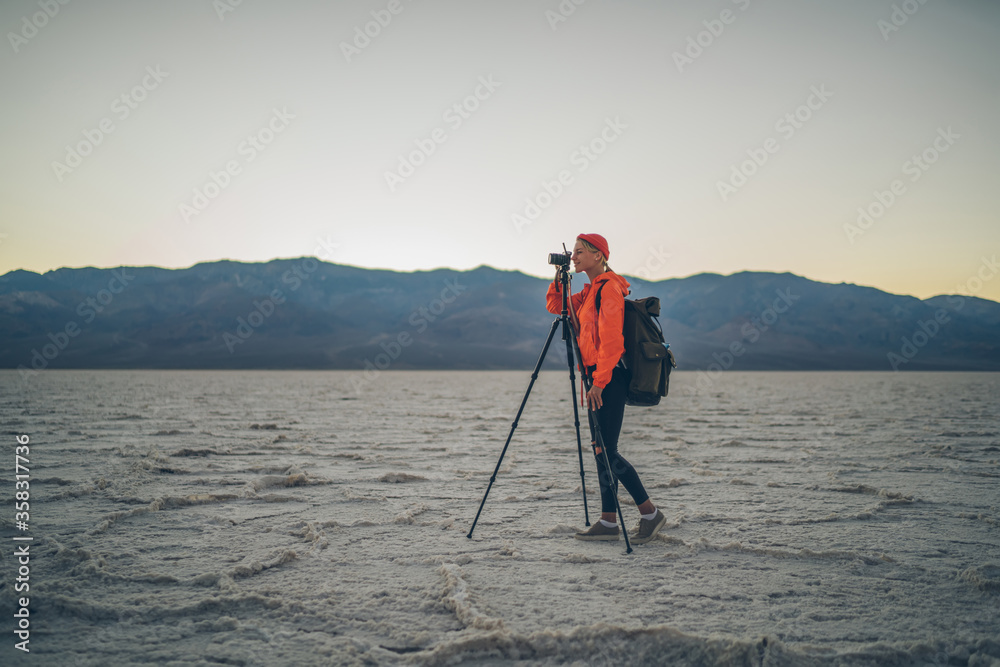 Woman traveler with backpack and in orange jaket taking video of landscape and sunset in desert using tripod,professional female photographer using modern equipment for making photo of wild nature