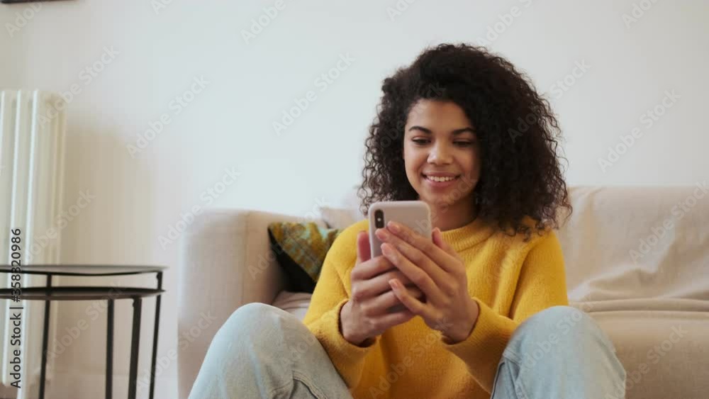 Joyful african attractive woman using smartphone while sitting on the floor in living room Joyful african attractive woman using smartphone while sitting on the floor in living room