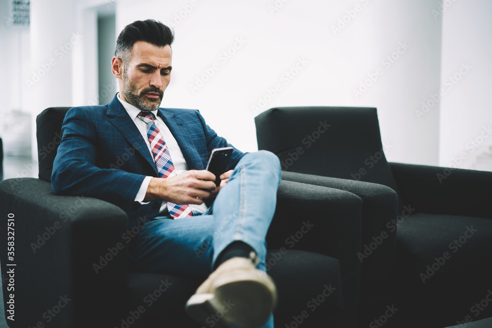 Pensive mature entrepreneur dressed in formal wear reading financial news on website on smartphone via 4G internet sitting on armchair in office interior.Concentrated proud ceo checking mail Pensive mature entrepreneur dressed in formal wear reading financial news on website on smartphone via 4G internet sitting on armchair in office interior.Concentrated proud ceo checking mail