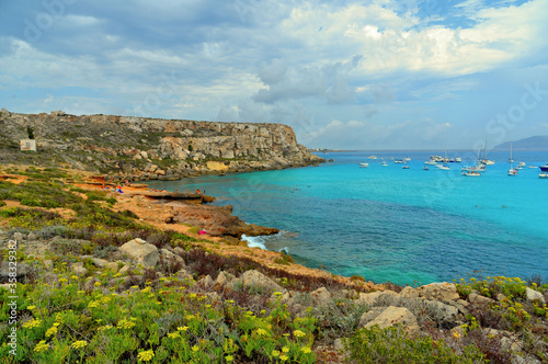 paradise clear torquoise blue water with boats and cloudy blue sky in background in Favignana island, Cala Rossa Beach, Sicily South Italy.