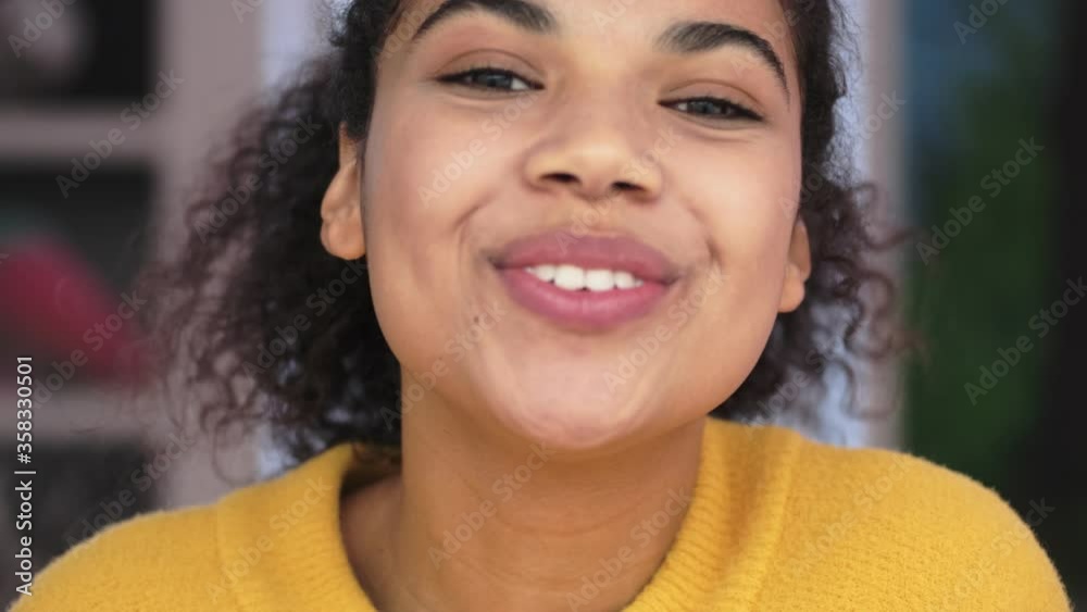 Close up view of Joyful african attractive woman in yellow sweater sending air kiss at the camera in living room
