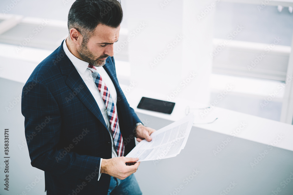 Top view of pensive mature entrepreneur dressed in formal wear reading financial reports with information data.Concentrated businessman 50 years old checking accounting text documents standing indoors Top view of pensive mature entrepreneur dressed in formal wear reading financial reports with information data.Concentrated businessman 50 years old checking accounting text documents standing indoors