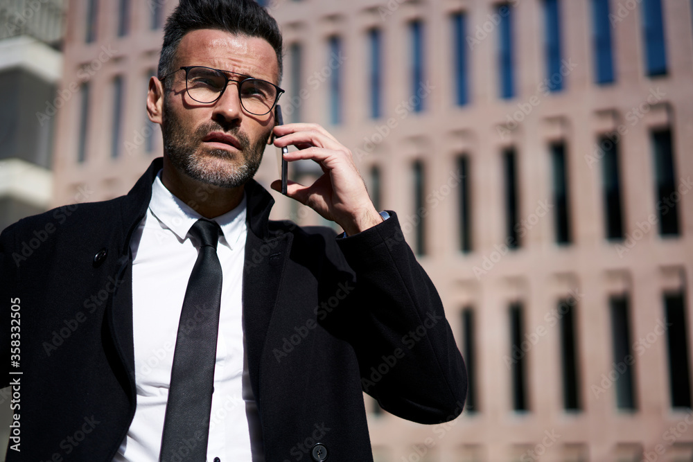 Successful bearded businessman or mature 40 years old executive with stylish glasses, black tie dressed and coat calling via modern telephone to employee. Copy space area for advertise text message