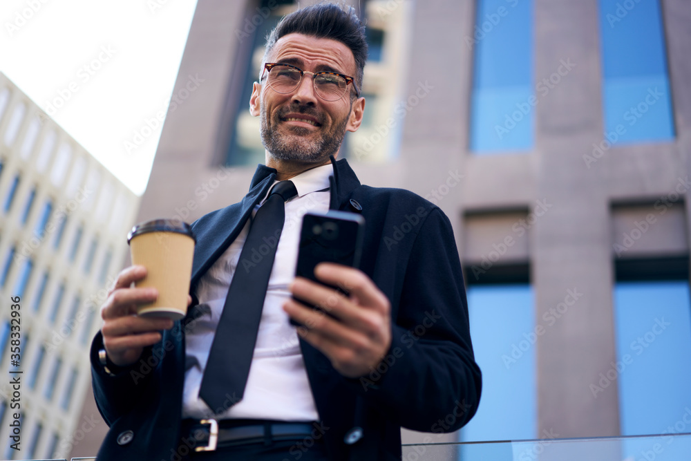 Fototapeta premium Positive good looking confident employer corporate dressed with mobile phone using high speed internet technology writing new message in social network to wife walking near big office building in city