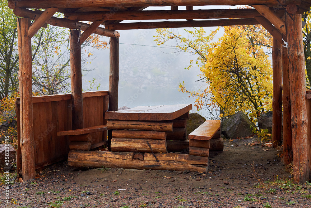 A gazebo with a wooden table near the lake in the early foggy autumn morning