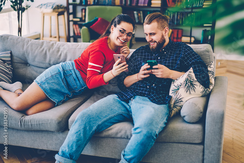 Cheerful romantic couple watching funny video from social networks on mobile phone resting on sofa, positive hipster girl showing common photos on smartphone to her boyfriend laughing together
