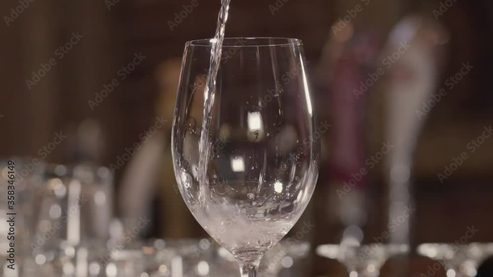 Static camera. Close up of a wine chalice in which is poured, from above, some white wine. Glass half full, other glasses in the blurred background. 