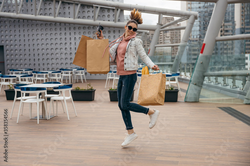 Happy young woman holding coffee cup, jumping with grocery shopping paper bag with long white bread baguette over city background