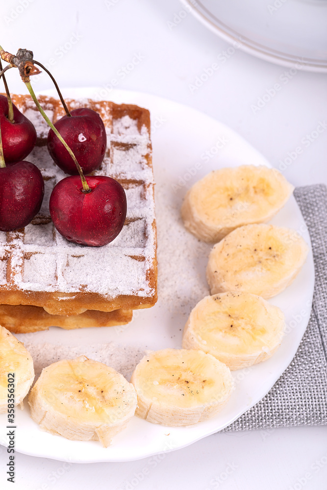 Belgian waffles in powdered sugar with bananas and cherries on a white plate. In the background, a teapot and a Cup for morning tea. Delicious Belgian waffles in powdered sugar with bananas and cherries on a white plate. In the background, a teapot and a Cup for morning tea. Delicious