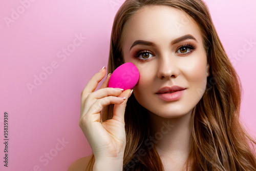 Portrait of a beautiful girl holding a beauty blender for makeup in the studio on a pink background