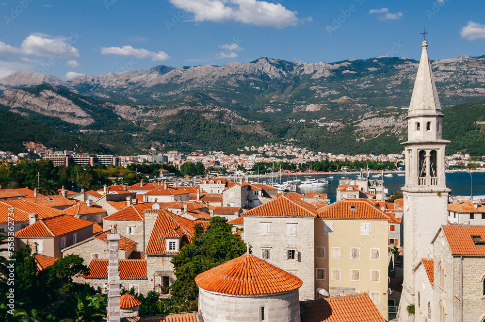 Landscape in Montenegro. Top view of Budva city. In the photo - old masonry buildings, cherished roofs, sharp roof spiers against the backdrop of green mountains and the blue Adriatic Sea with boats