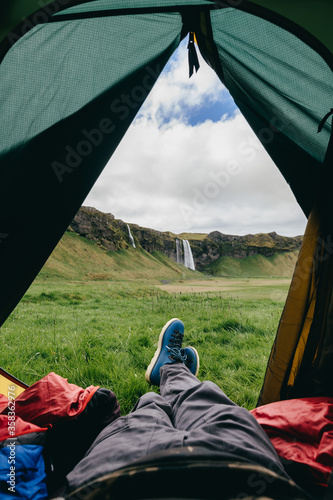 Icelandic waterfall view from the green tourist tent among endless bright fields. A photographer rests in a tent and takes a photograph from a lying position. 