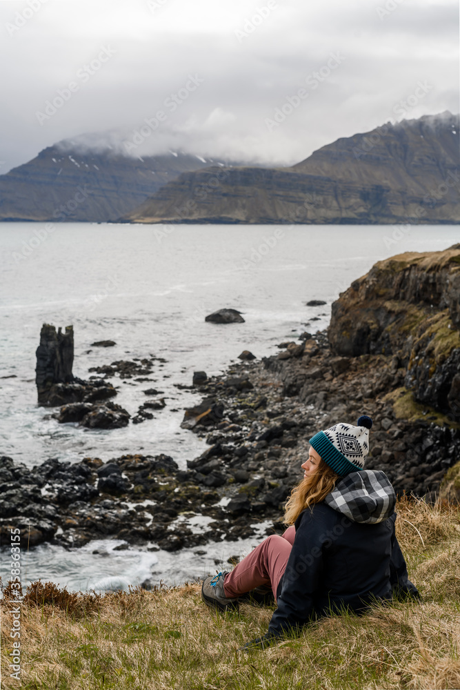Fototapeta premium A beautiful girl with long blonde hair and in warm clothes is sitting on the cliffed coast of the Atlantic Ocean. She admires the Icelandic landscape on the background of a volcanic beach