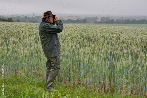 A hunter stands by a wheat field and observes his hunting area through binoculars on a rainy, foggy morning in June. In the background you can see the small town of Trossingen in Germany. © BIB-Bilder