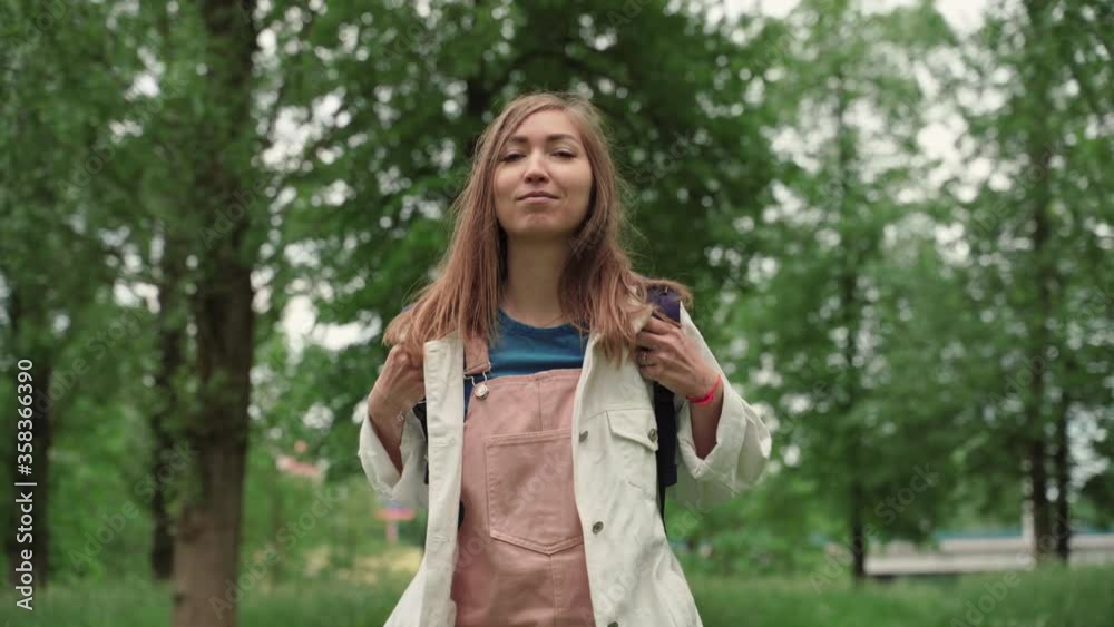 Beautiful woman smiles walking along path surrounded by foliage and trees. Front view of female with backpack walking at the summer park. Beautiful woman smiles walking along path surrounded by foliage and trees. Front view of female with backpack walking at the summer park.